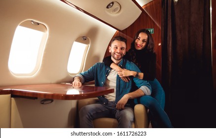 Our Happy Family. A Lovely Couple Is Sitting Together On An Armchair In A Private Jet, Hugging And Smiling Out Of Joy.