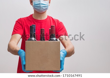  ourier wearing protective mask and gloves holding box with bottles of wine. Safe online wine store delivery during quarantine.