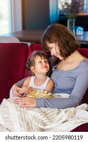 Out Of Focus Background Of Mom And Daughter Are Sitting On A Bright Sofa And Hugging. They Are Covered With A Warm Blanket And Eat Popcorn Together. The Concept Of Love And Comfort Family