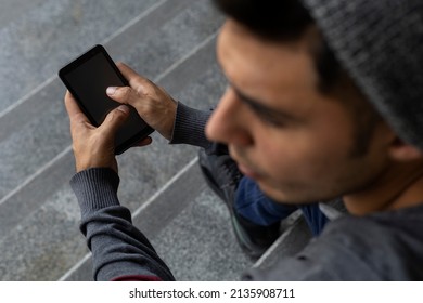 Out Of Focus Profile Of Young Male (22) Latin American Sitting On The Stairs Checking His Cell Phone. View From Above. Technology Concept.