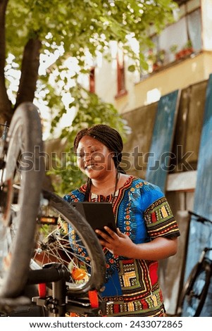 At outdoor active vibrant black woman exploring the internet for bike maintenance. Female african american cyclist thoroughly inspecting and repairing bicycle with digital tablet.