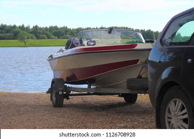 Outdoor activities, boat launch - descent of a white-red motor plastic boat on a trailer with a car on the water on a sandy slipway on the background of the river and the shore with green trees