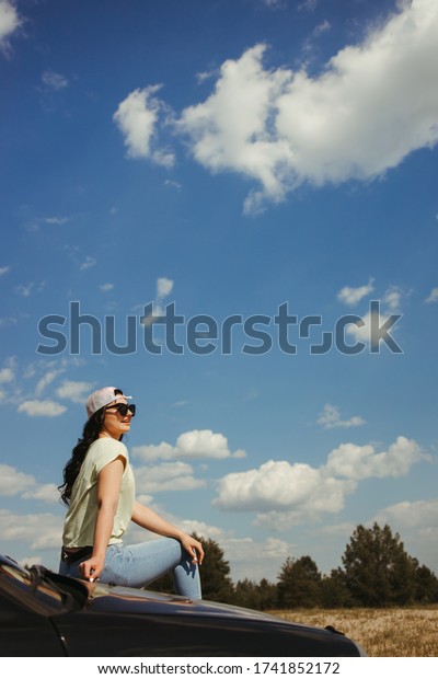 Outdoor adventure. Young woman sitting on the car\
hood while traveling. Tourism lifestyle, vacation, freedom and\
travel concept