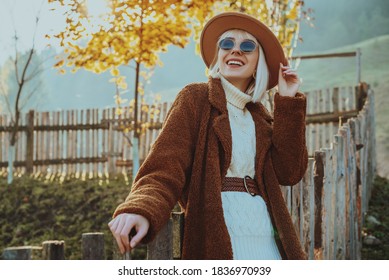 Outdoor autumn fashion, lifestyle portrait of smiling woman, model wearing trendy outfit with hat, sunglasses, faux fur coat, knitted sweater, wicker belt, posing on nature. Copy, empty space for text