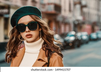Outdoor autumn portrait of young elegant fashionable lady wearing trendy big sunglasses, green beret, hoop earrings, camel coat, white turtleneck,walking in street of European city. Copy, empty space
