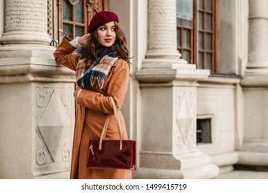 Outdoor autumn portrait of young elegant fashionable lady wearing trendy beret, camel coat, plaid scarf, holding dark red baguette bag, posing in street of European city. Copy, empty space for text