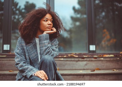 Outdoor autumn portrait of young thoughtful african american woman looking aside sitting on old wooden house stairs, pensive curly girl spending time outside on fall day, thinking about life - Powered by Shutterstock