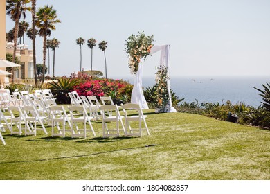 Outdoor beach ceremony area with ocean view, white chairs, floral arch on sunny day
