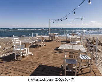 Outdoor beachfront cafe with tables and chairs under a wooden pergola overlooking the sea with palm trees and sun loungers nearby - Powered by Shutterstock