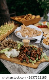 An outdoor buffet table with assorted appetizers including meat skewers, bread, and vegetables on a rustic wooden table.