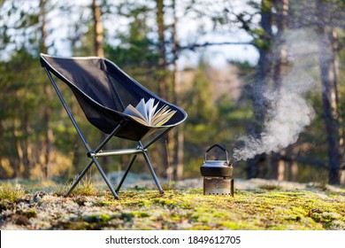 Outdoor Camping Equipment Close-up. Stove With Kettle, Tourist Folding Chair With A Book In The Forest. The Background Is Blurred.