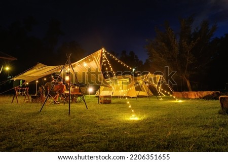 Outdoor camping tent with tarp or flysheet on grass courtyard and warm night light under dark night sky, family vacation picnic on holiday relax.