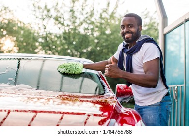 Outdoor Car Wash With Sponge And Foam. Car Washing. Smiling African Man Showing Thumb Up, Cleaning His Car Windscreen, Using Sponge And Foam In The Outdoor Car Wash Station