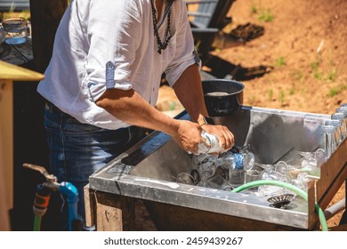 Outdoor clean: man in white shirt and jeans diligently washing dishes in an open-air sink - Powered by Shutterstock