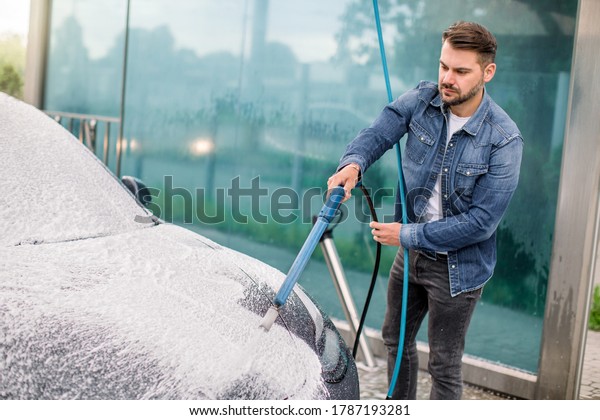 Outdoor\
cleaning of the car with soap foam using high pressure jet.\
Handsome bearded young man washing his electric car under high\
pressure with foam in self wash service\
outdoors.