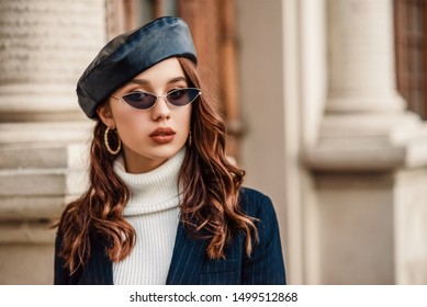 Outdoor close up autumn portrait of young elegant fashionable woman wearing trendy leather beret, sunglasses, hoop earrings, blazer, turtleneck, posing in street of European city. Copy, empty space 