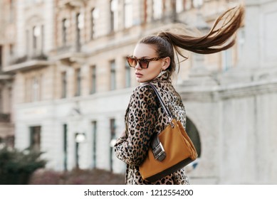 Outdoor close up fashion portrait of young beautiful fashionable woman wearing  sunglasses, leopard print blazer, holding brown suede bag, walking in street of european city. Copy, empty space