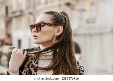 Outdoor close up fashion portrait of young beautiful fashionable woman wearing stylish animal, leopard print sunglasses, hoop earrings, turtleneck, posing in street of european city. Copy, empty space