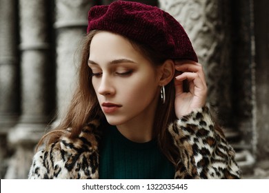 Outdoor close up fashion portrait of young beautiful fashionable lady wearing trendy animal, leopard print faux fur coat, beret, hoop earrings, posing in street of  European city