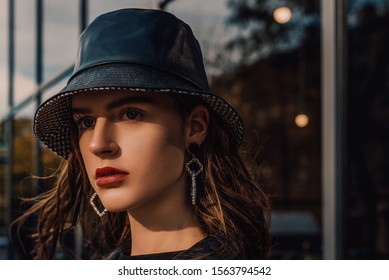Outdoor Close Up Fashion Portrait Of Young Beautiful Confident Model, Woman Wearing Trendy Faux Leather Bucket Hat, Long Earrings, Trench Coat, Posing In City Street. Copy, Empty Space For Text