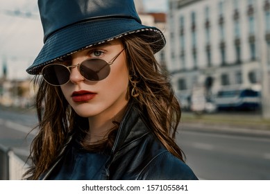 Outdoor Close Up Fashion Portrait Of Young Elegant Model, Woman Wearing Trendy Faux Leather Bucket Hat, Round Sunglasses, Earrings, Posing In Street Of European City. Copy, Empty Space For Text