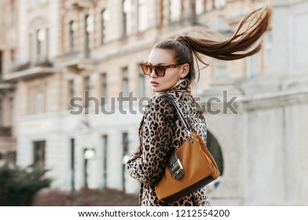 Outdoor close up fashion portrait of young beautiful fashionable woman wearing  sunglasses, leopard print blazer, holding brown suede bag, walking in street of european city. Copy, empty space