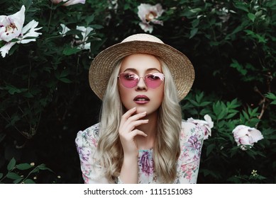 Outdoor close up portrait of beautiful young woman with long blonde hair, makeup, wearing pink cat eye sunglasses, straw boater hat posing in the blooming garden.  Female spring fashion concept