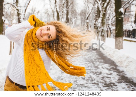 Outdoor close up portrait of blond curly playful hipster woman on street, looking at camera, smiling at snowy park. Model wearing white sweater, yelow winter hat, sarf, gloves. City lifestyle