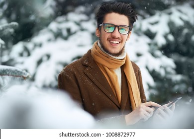 Outdoor Close Up Portrait Of Young Beautiful Men. Smiling Young Man Having Fun Outdoors. Businessman Uses A Phone . Man Enjoy A Winter. Dressed In A Coat, Sweater And Scarf. Winter Concept. Snowfall.