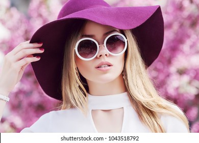 Outdoor close up portrait of young beautiful girl wearing stylish round sunglasses, trendy violet hat, white shirt, posing in street. Pink blooming trees on background. Spring fashion concept. 