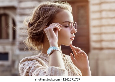 Outdoor close up portrait of young beautiful fashionable girl wearing purple sunglasses, sweater, wrist watch, posing in street of european city. Copy, empty space for text