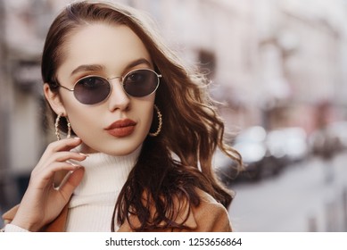 Outdoor close up portrait of young beautiful woman with long hair wearing  stylish sunglasses, white turtleneck, coat, model posing in street of european city. Copy, empty space for text