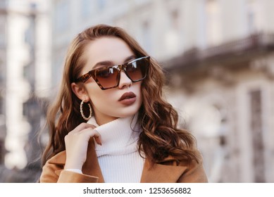 Outdoor close up portrait of young beautiful woman wearing trendy leopard print sunglasses, hoop earrings,  white turtleneck, coat, model posing in street of european city. Copy, empty space for text