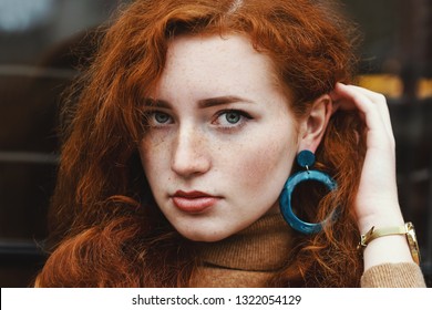 Outdoor close up portrait of young beautiful redhead lady with natural long curly hair and feckled skin, wearing big blue plastic circle earrings, looking at camera, posing in street