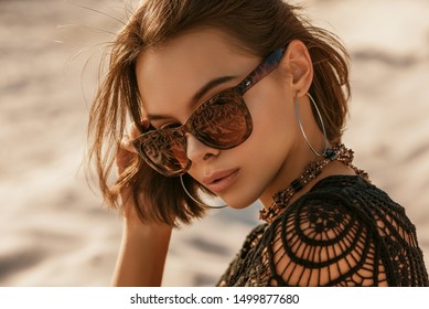 Outdoor close up portrait of young beautiful woman with tanned skin wearing luxury wide frame square sunglasses, hoop earrings, crochet top, posing on sand, at sunset. Copy, empty space for text 