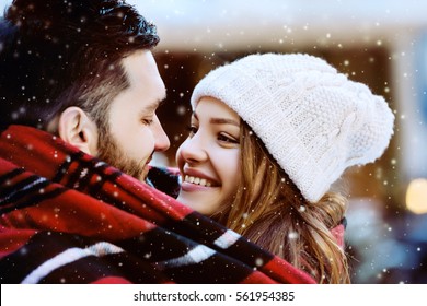 Outdoor Close Up Portrait Of Young Beautiful Happy Smiling Couple. Models Looking At Each Other, Bundled Up In Tartan Blanket. Christmas, New Year, Winter Holidays, Dating Concept. Day Light, Snowfall