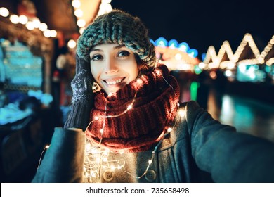 Outdoor close up portrait of young beautiful happy smiling girl making selfie photo in night street. Festive Christmas fair on background. Model looking at camera, wearing knitted beanie hat, scarf.