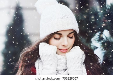 Outdoor close up portrait of young beautiful happy smiling girl wearing white knitted beanie hat, scarf and gloves. Model posing in street. Winter holidays concept. Toned