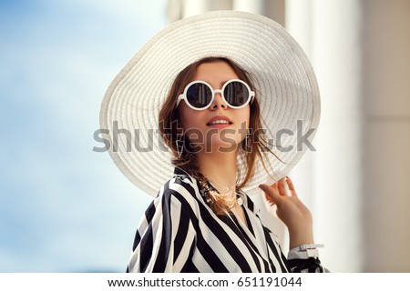 Outdoor close up portrait of young beautiful happy girl posing in street. Model wearing stylish round sunglasses, wide-brimmed hat, stripped black-white blouse. City lifestyle, female fashion concept
