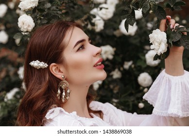 Outdoor close up portrait of young fashionable happy smiling lady wearing trendy vintage style earrings, white pearl barrette in the long hair, posing in the blooming rose garden - Shutterstock ID 1438869038