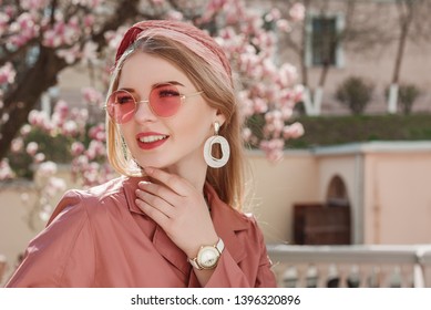 Outdoor close up spring fashion portrait of young beautiful happy smiling lady wearing trendy earrings, wrist  watch, stylish pink round sunglasses, headband, coat, posing in street. Copy, empty space