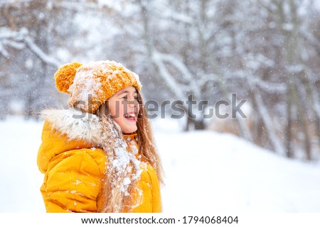 Outdoor close-up portrait of young beautiful happy smiling girl, wearing yellow jacket and knitted hat walking in winter park
