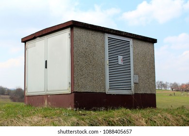 Outdoor concrete electrical high voltage box with two metal doors and one ventilation side opening surrounded with high uncut grass and cloudy sky in background - Shutterstock ID 1088405657