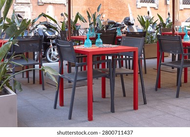 Outdoor dining setup features red tables and black chairs, surrounded by plants and parked motorcycles in a vibrant urban setting