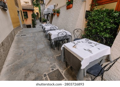 Outdoor dining setup features tables with white tablecloths and cutlery along a cobblestone street in Varenna, surrounded by greenery