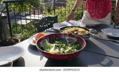 Outdoor dining table set for a meal with colorful salad, pasta, and fresh fruit slices. A person stands by the table, adding a touch of elegance to the cozy garden setting - Powered by Shutterstock