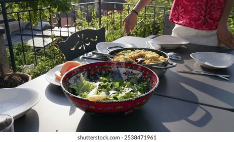 Outdoor dining table set for a meal with colorful salad, pasta, and fresh fruit slices. A person stands by the table, adding a touch of elegance to the cozy garden setting - Powered by Shutterstock