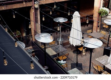 Outdoor Dining Tables At A Restaurant With String Lights Hanging Above Located In A Shopping Plaza.  