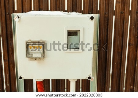 Outdoor electrical control box with meter mounted on wooden fence covered by snow in winter sunlight