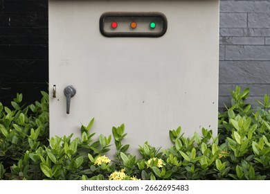 Outdoor electrical control panel with indicator lights (red, orange, green) and a handle, surrounded by green foliage.